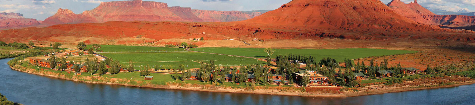 The image features a scenic landscape with a river curving in the foreground, lush green fields, buildings, and a backdrop of red rock formations.