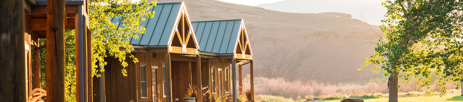 The image shows a scenic outdoors with small wood cabins, a curved path, lush green lawn, trees, and mountains in the background under soft sunlight.