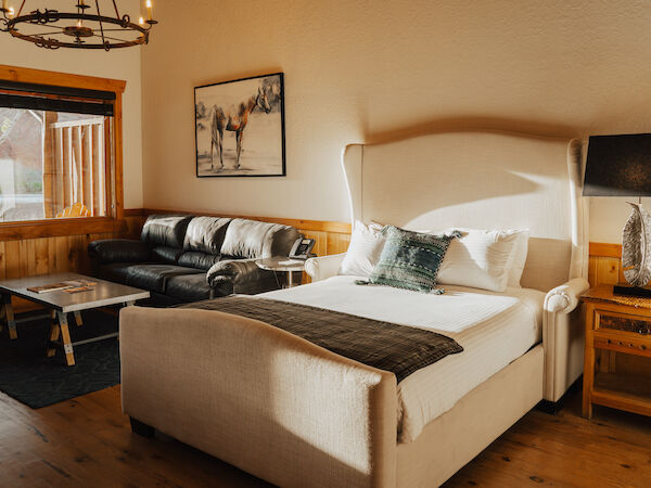 A cozy bedroom with a large bed, sofa, and wooden furniture. A wall painting and a lamp add charm to the warm, sunlit space.