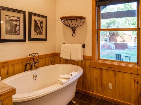 The image shows a cozy bathroom with a freestanding tub, two framed pictures, towels, wooden paneling, and a window with an outdoor view.