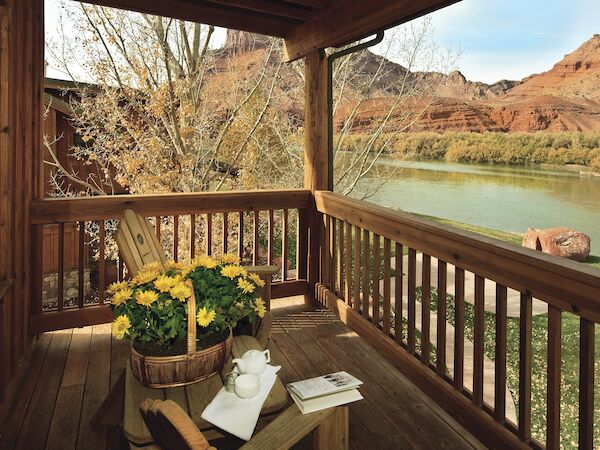 A wooden porch overlooks a river and mountains, featuring a table with flowers, an open book, and a chair with yellow cushions.