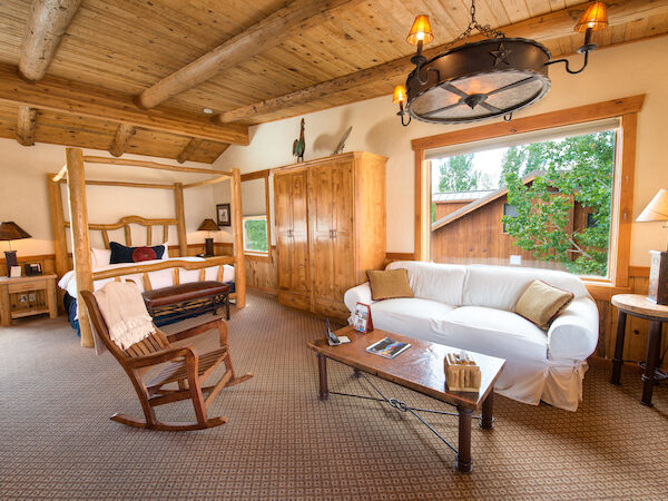 A cozy rustic room with wooden ceiling, a bed, a sofa, a rocking chair, and a coffee table. Large windows show greenery outside.