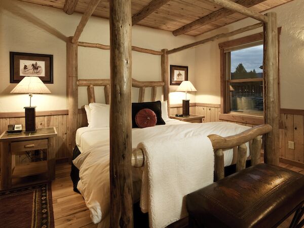 A cozy bedroom with a rustic wooden canopy bed, two nightstands with lamps, framed pictures, and a bench at the foot of the bed.