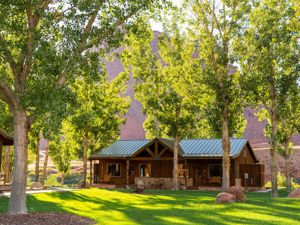A rustic cabin surrounded by lush trees, sitting on a manicured lawn with rocky hills in the background, bathed in warm sunlight.