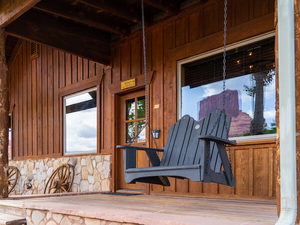 A wooden porch features a hanging swing, rustic decor, and windows reflecting a scenic view of a distant butte under a cloudy sky.
