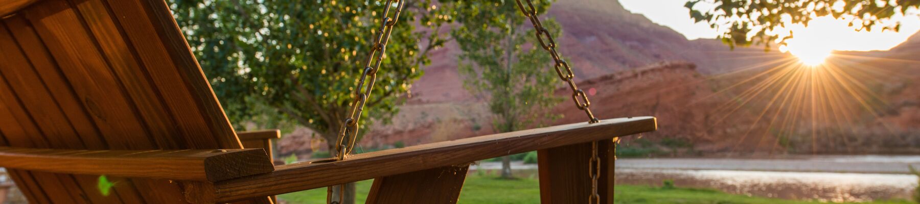 A wooden swing on a porch overlooks a scenic landscape with a river, trees, and a mountain as the sun sets in the distance.