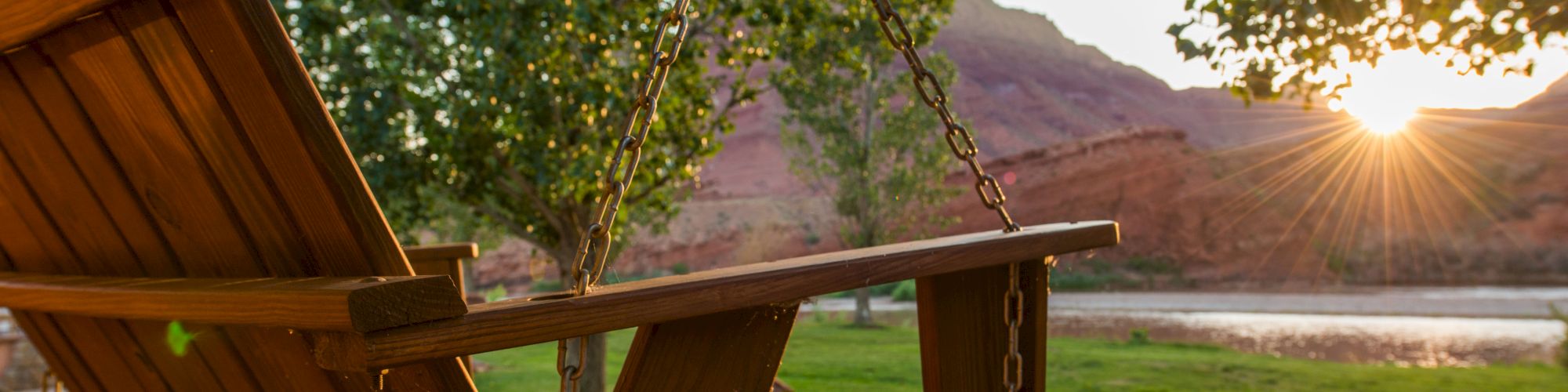 A wooden swing on a porch overlooks a scenic landscape with a river, trees, and a mountain as the sun sets in the distance.
