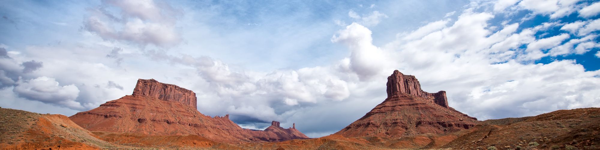 The image shows a desert landscape with red rock formations and buttes under a partly cloudy sky. The terrain is mostly barren with sparse vegetation.
