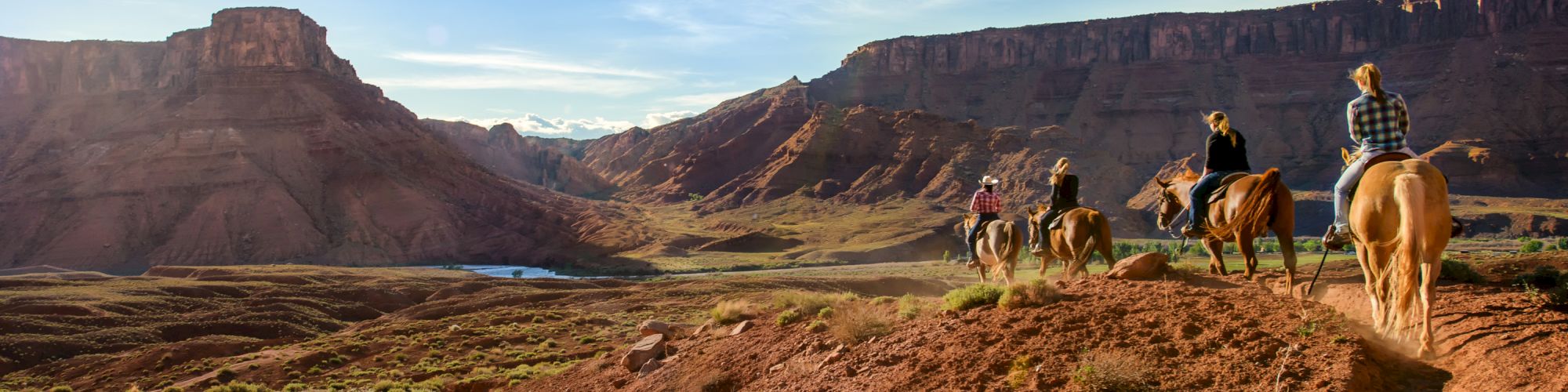 The image shows a group of people riding horses on a trail through a scenic canyon landscape with blue skies and sunlight illuminating the area.
