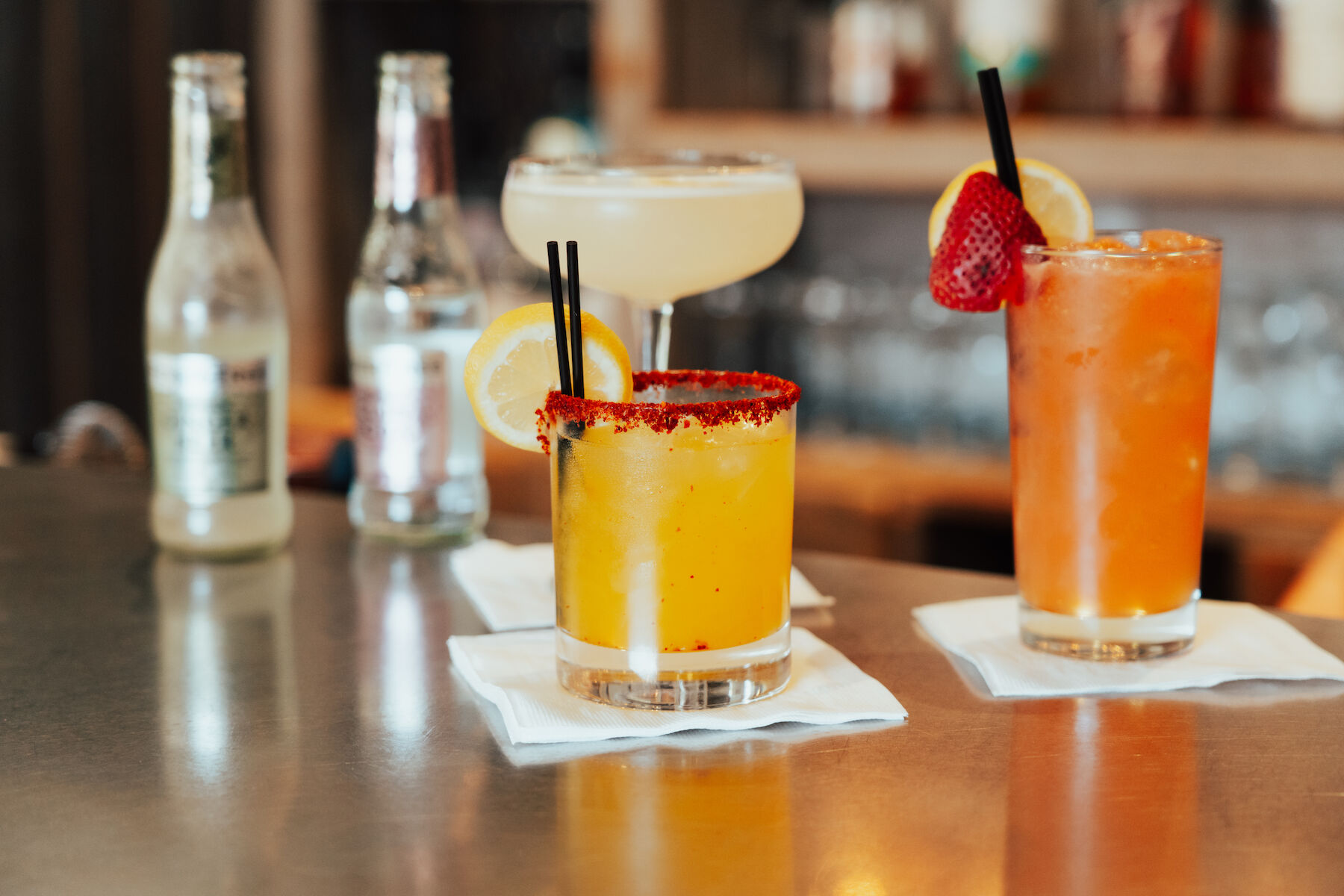 Various cocktails on a bar counter, garnished with fruit. Two bottles are in the background on the left.