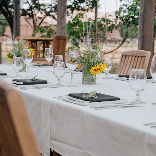 An outdoor dining setup with a white tablecloth, wine glasses, black napkins, and floral centerpieces, surrounded by wooden chairs.