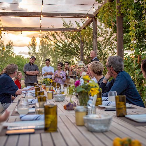 A group of people is gathered around a long outdoor dining table with string lights above, enjoying an evening meal under a pergola with greenery.