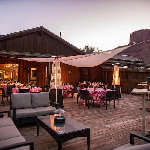An outdoor restaurant patio with seating, heaters, and a wooden deck. Tables are set with checkered tablecloths, surrounded by a scenic mountain backdrop.