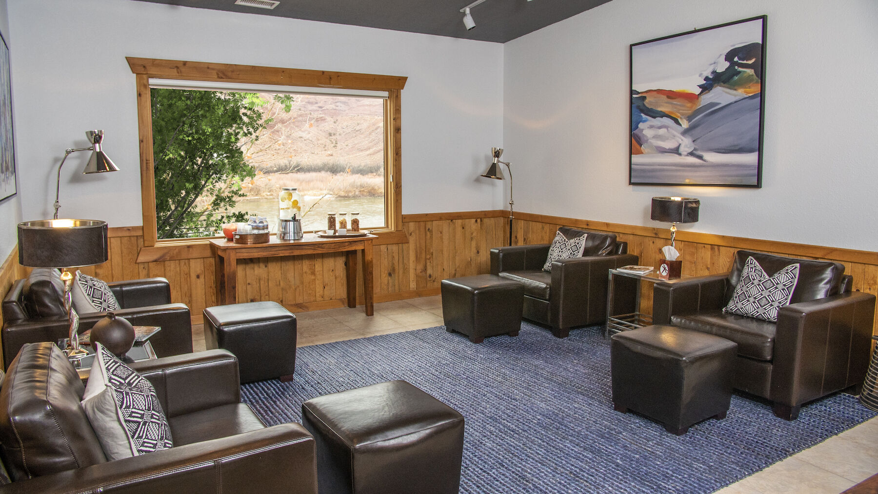 The image shows a cozy lounge with leather chairs, wooden paneling, a large window view, a table with refreshments, ambient lighting, and a stylish rug.