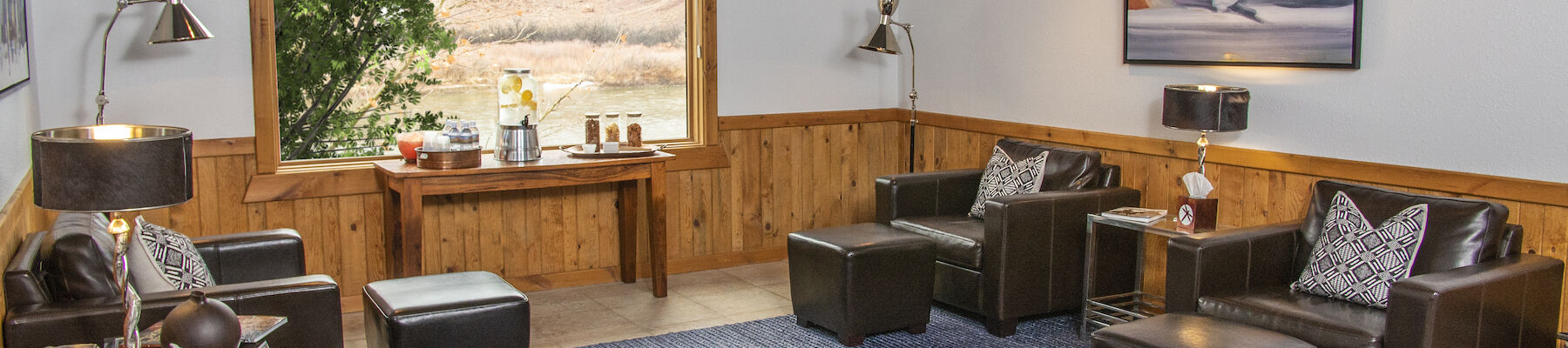 The image shows a cozy lounge with leather chairs, wooden paneling, a large window view, a table with refreshments, ambient lighting, and a stylish rug.