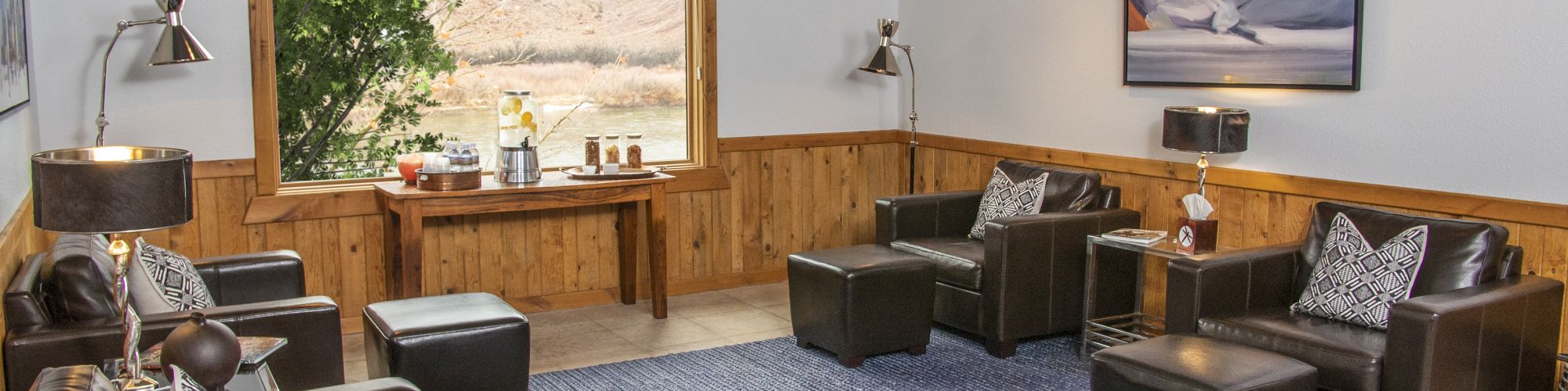 The image shows a cozy lounge with leather chairs, wooden paneling, a large window view, a table with refreshments, ambient lighting, and a stylish rug.