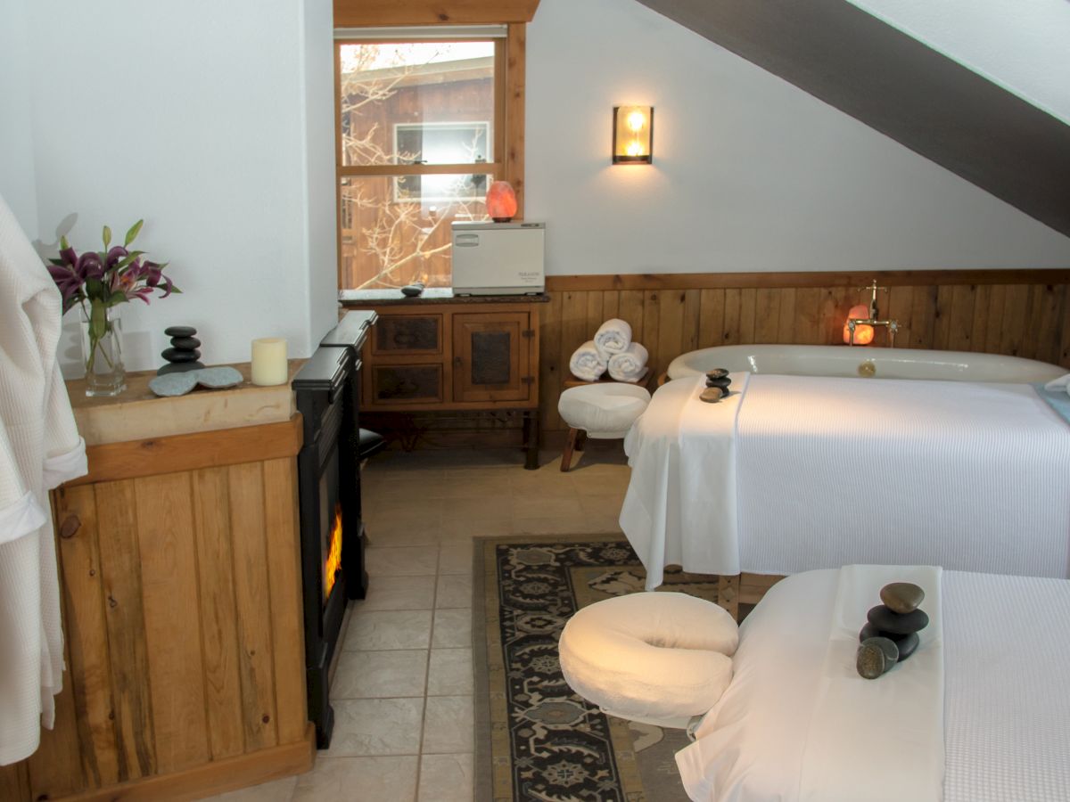 A cozy spa room with two massage tables, neatly folded towels, a cosy fireplace, and plush bathrobes creating a relaxing atmosphere.