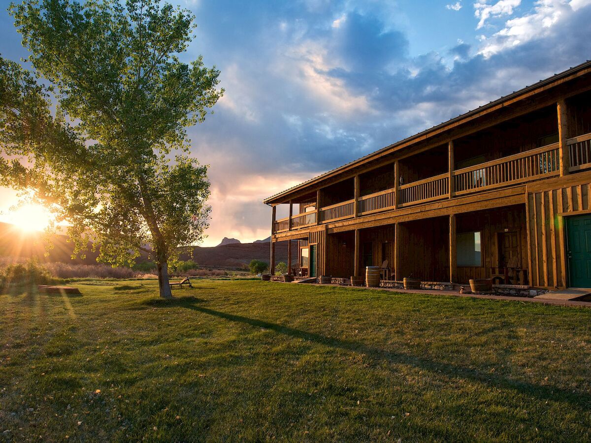 A wooden building stands beside a tree at sunset, casting long shadows on the grass with a partly cloudy sky in the background.