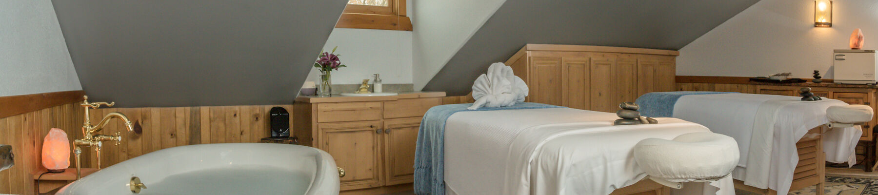 This image shows a cozy spa room with two massage tables, a bathtub, wooden furniture, and soft lighting providing a relaxing ambiance.