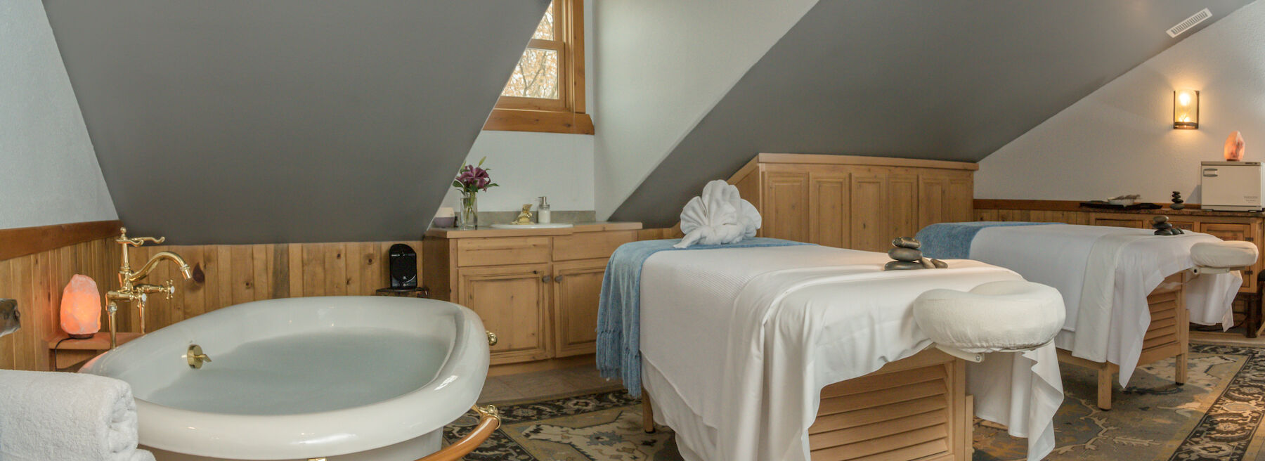This image shows a cozy spa room with two massage tables, a bathtub, wooden furniture, and soft lighting providing a relaxing ambiance.