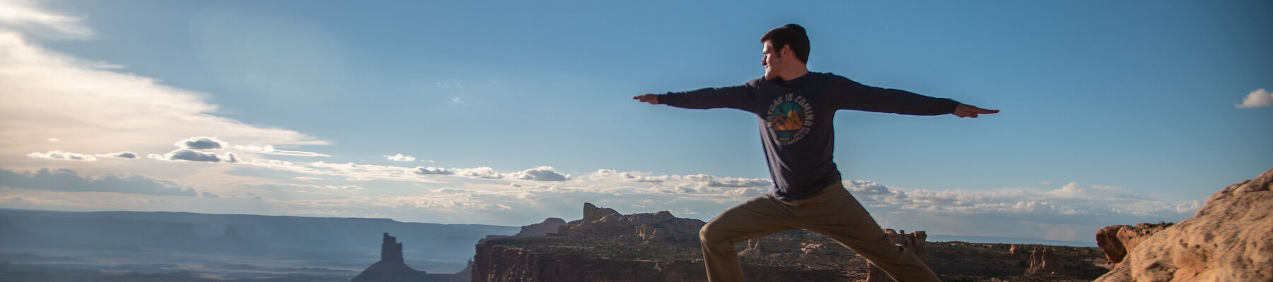 A person performs a yoga pose on a rocky ledge with a stunning, expansive desert landscape and distant rock formations under a clear blue sky.