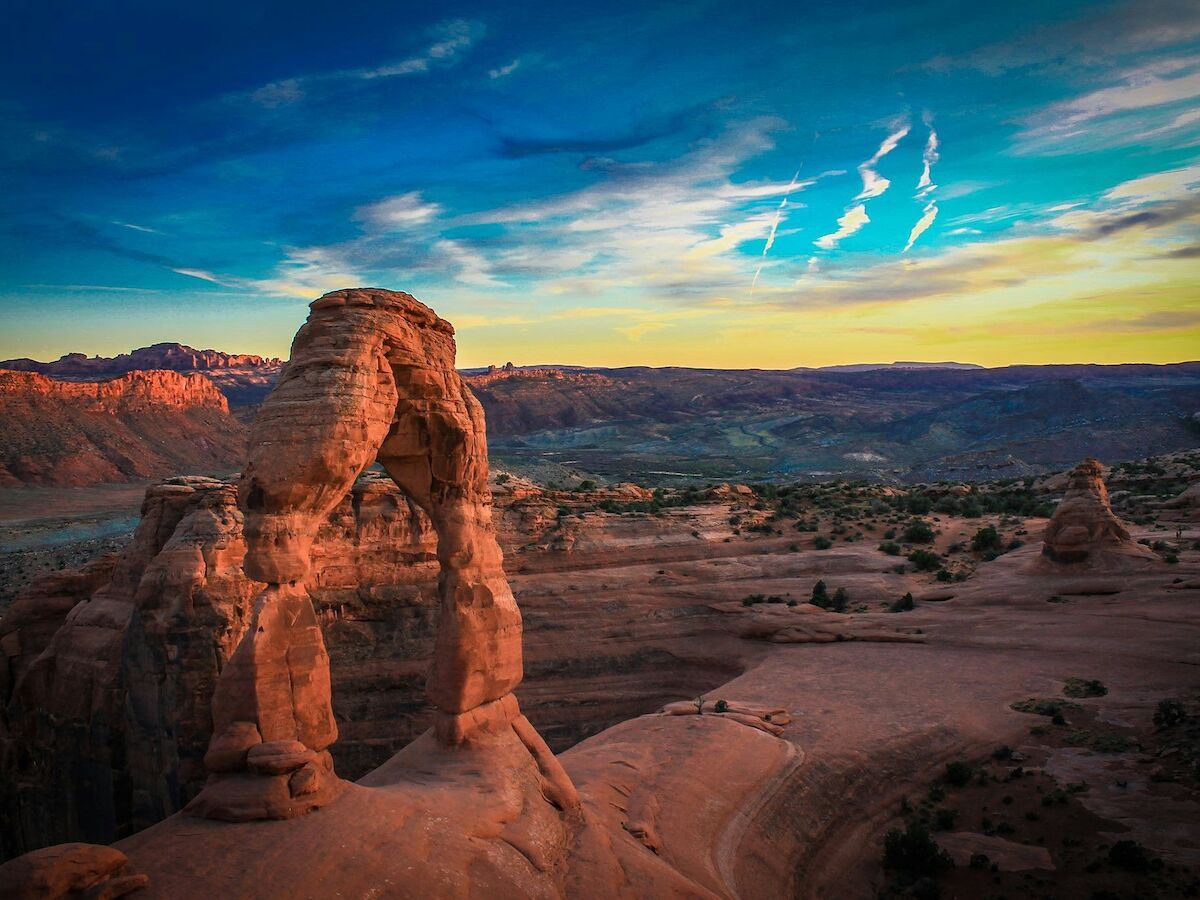 A scenic view of Delicate Arch in Arches National Park at sunset with vibrant skies and colorful rock formations in the background.