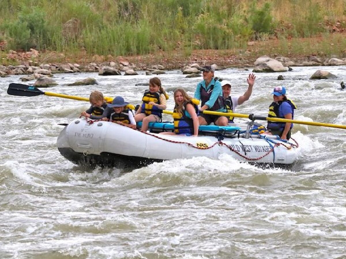 A group of people wearing life jackets are white water rafting on a river, all looking engaged and one person is waving towards the camera.