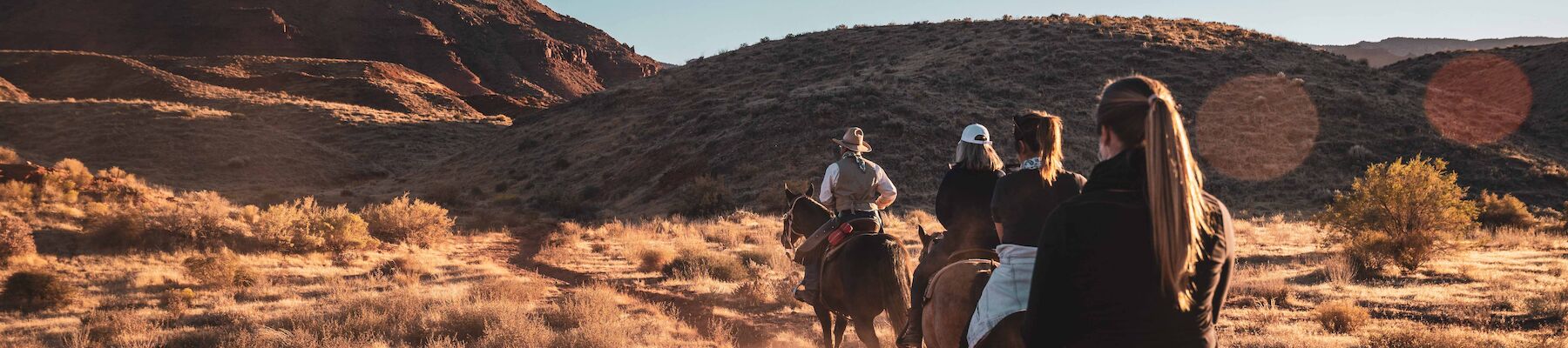 People riding horses on a dusty trail in a desert landscape with a large rock formation in the background under a clear sky.