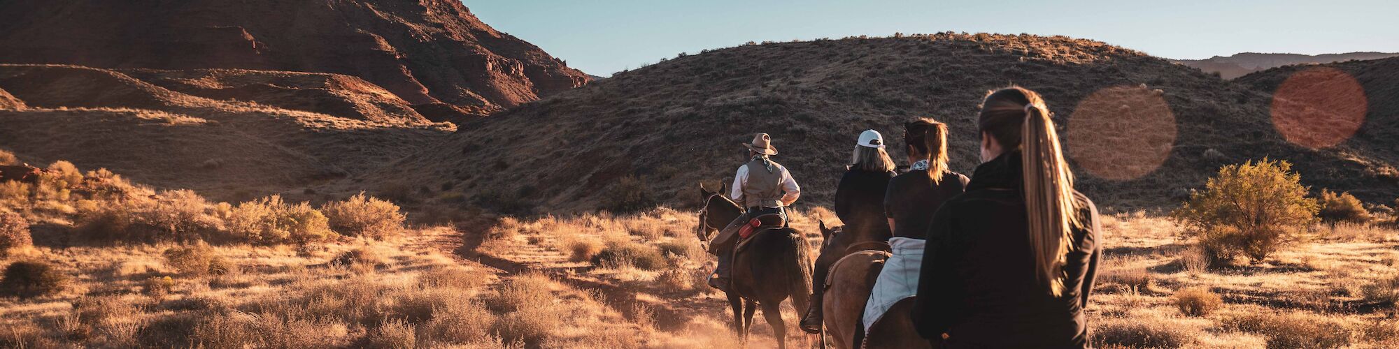 People riding horses on a dusty trail in a desert landscape with a large rock formation in the background under a clear sky.