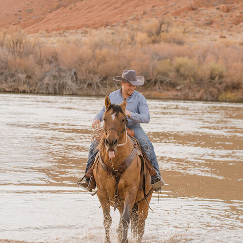 A person wearing a hat is riding a horse through a shallow river, with a rocky landscape in the background.