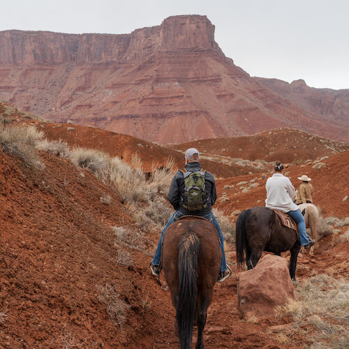 Three people riding horses through a rugged, red desert landscape with large rock formations in the background.