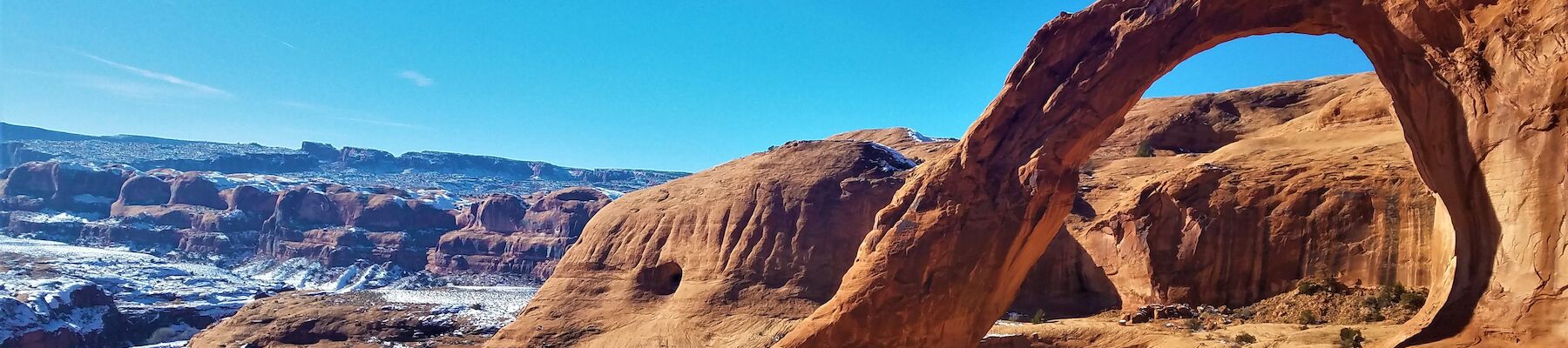 A natural sandstone arch in a desert landscape with clear blue skies and distant snow-covered areas is captured in the image.