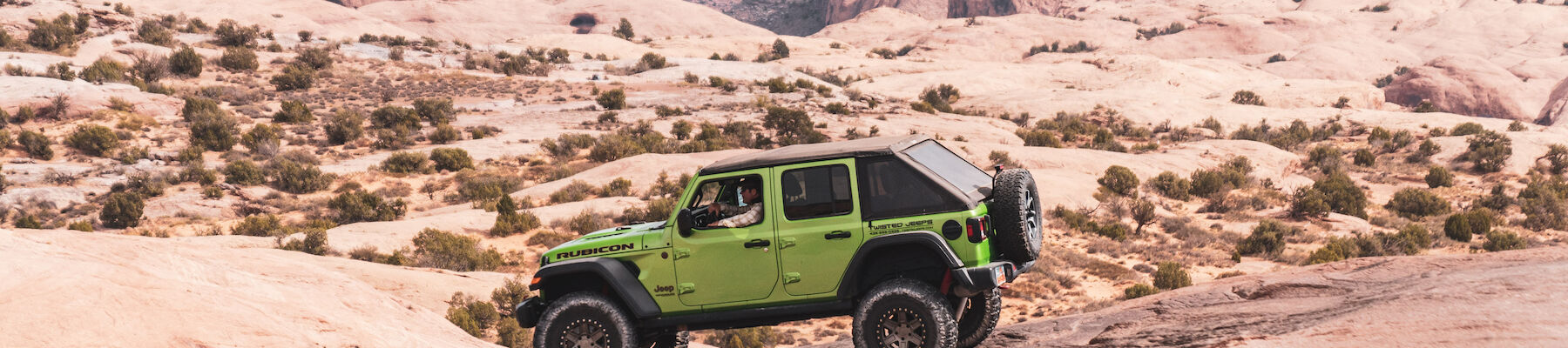 A green off-road vehicle traverses a rocky desert landscape with distant hills and canyons in the background.