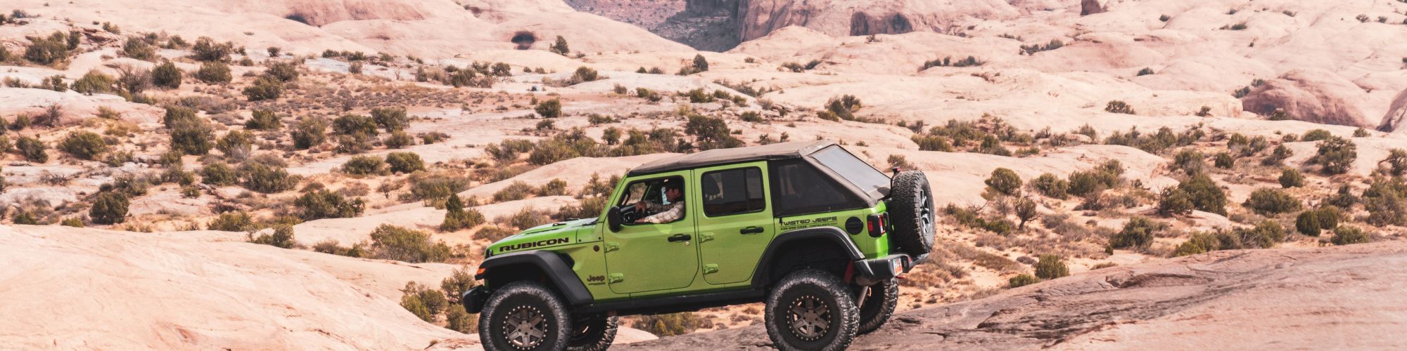 A green off-road vehicle traverses a rocky desert landscape with distant hills and canyons in the background.