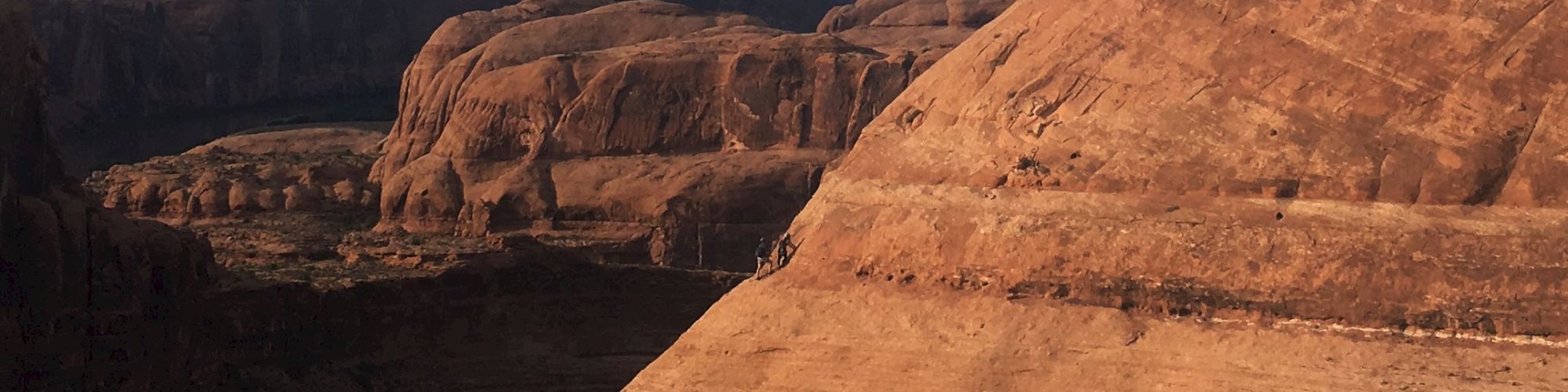 The image shows a person standing atop a large, reddish rock formation in a rugged, mountainous landscape under a blue sky with scattered clouds.