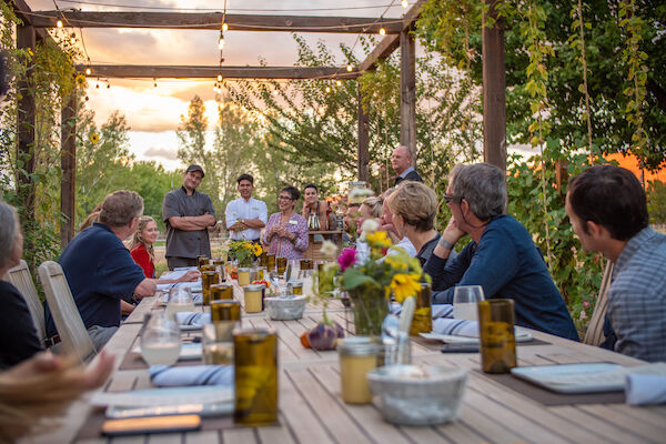 A group of people is gathered around a long outdoor dining table decorated with flowers, while others stand and make a toast under string lights at sunset.