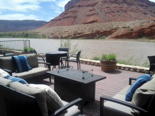A cozy outdoor seating area with sofas, chairs, and a table overlooking a river and red rock cliffs under a blue sky.
