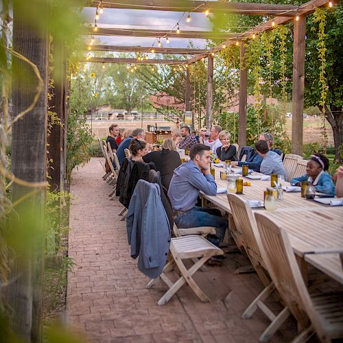 A group of people are seated at a long outdoor table under a pergola with string lights, enjoying a meal. The setting appears to be cozy and rustic.