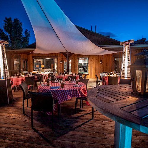 An outdoor dining area is set up with tables covered in checkered tablecloths and lit by heaters and lanterns, creating a cozy atmosphere at dusk.