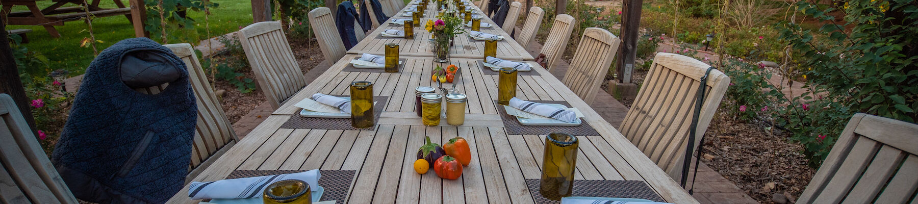 A long outdoor dining table set with plates, napkins, glasses, and small decorations, surrounded by wooden chairs in a garden setting.