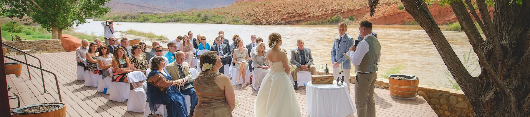 An outdoor wedding ceremony is taking place on a wooden deck, with a river and red rock landscape in the background.