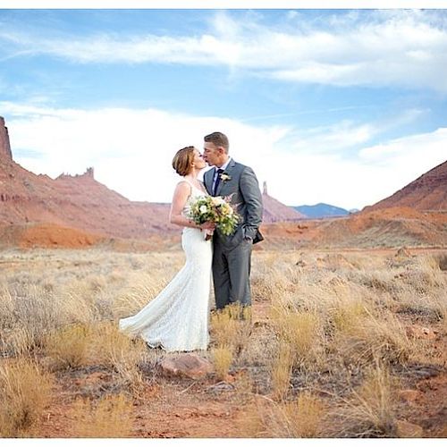A bride and groom kiss while standing in an open desert landscape with red rock formations in the background, holding a bouquet of flowers.