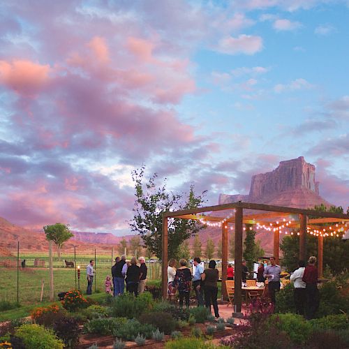 People are gathered under a lit wooden pergola in a garden with scenic mountains and a colorful sky in the background.