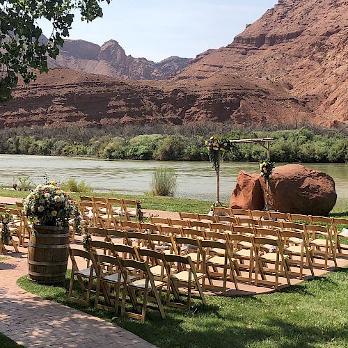 An outdoor wedding setup by a river with rows of wooden chairs, floral decorations, and a scenic backdrop of red rock formations, ending the sentence.