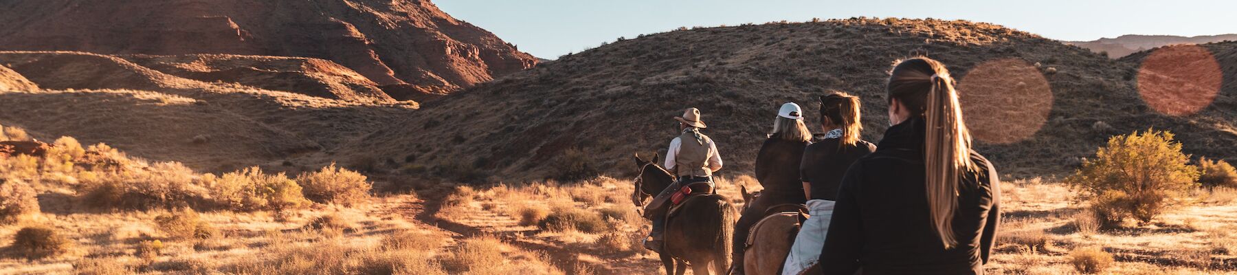 People riding horses across a dry, open landscape with a rocky mesa in the background under a clear blue sky.
