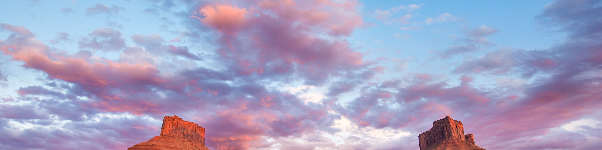 A vast desert landscape features two mesas under a colorful sky with pink and blue clouds during sunset.