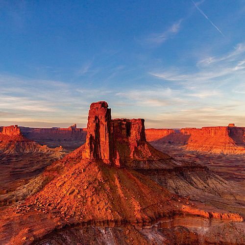 The image shows a towering red rock formation in a vast desert landscape under a blue sky with wispy clouds and a small moon visible above.