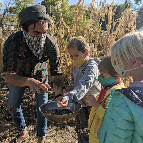 An adult and three children, all wearing masks, examine a bowl of seeds in a garden or farm setting.