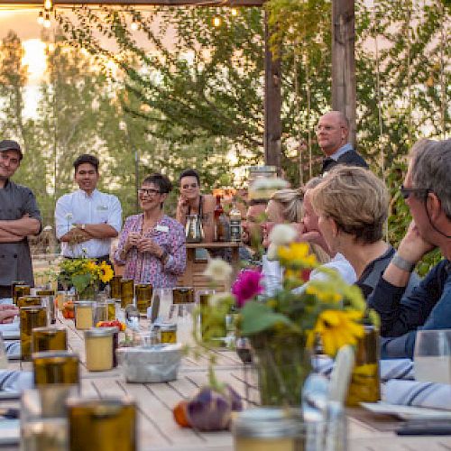 A group of people are gathered around a long table outdoors, enjoying a meal. Some are standing and appear to be speaking to the seated guests.