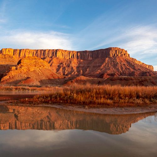 A rugged desert landscape with orange rock formations is reflected in a calm river under a clear, blue sky with scattered clouds.